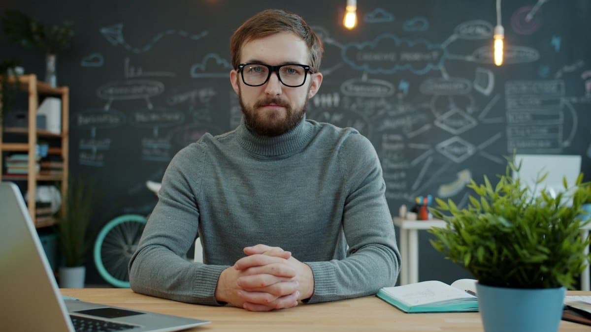 Man in glasses sitting at a desk in an office with a chalkboard behind him